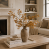 A rustic brown ceramic vase filled with dried golden-orange flowers, displayed on a stone tray atop a wooden coffee table in a cozy living room with a fireplace and sofa in the background.