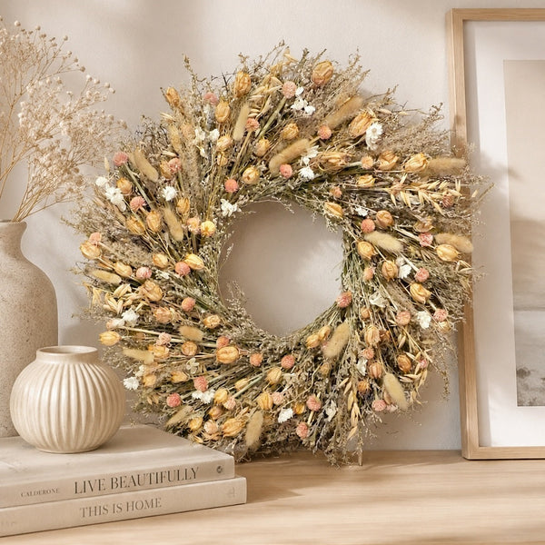 A round dried floral wreath in beige, tan, cream, and soft pink tones, displayed on a light wood console table beside books, a ribbed ceramic vase, and a framed print against a warm beige wall.