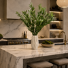 A modern kitchen island with a white marble countertop and matching marble backsplash, styled with a large green leafy arrangement in a textured white vase and a bowl of green fruit. The background features dark wood cabinetry, built-in shelves with warm lighting, and a gold faucet.