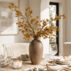 A rustic brown ceramic vase filled with delicate yellow-orange dried flowers, styled on a dining table with white plates, gold cutlery, and wine glasses in a warm, sunlit room.