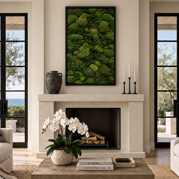 A framed vertical wall art piece featuring a dense green moss or pine forest texture hangs above a fireplace in a modern living room. The room has cream walls, black-framed windows, a light stone fireplace, a wooden coffee table with white orchids, and candles on a side table.