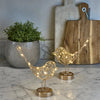 Decorative gold bird sculptures with string lights on a marble surface, with a plant and cutting board in the background.
