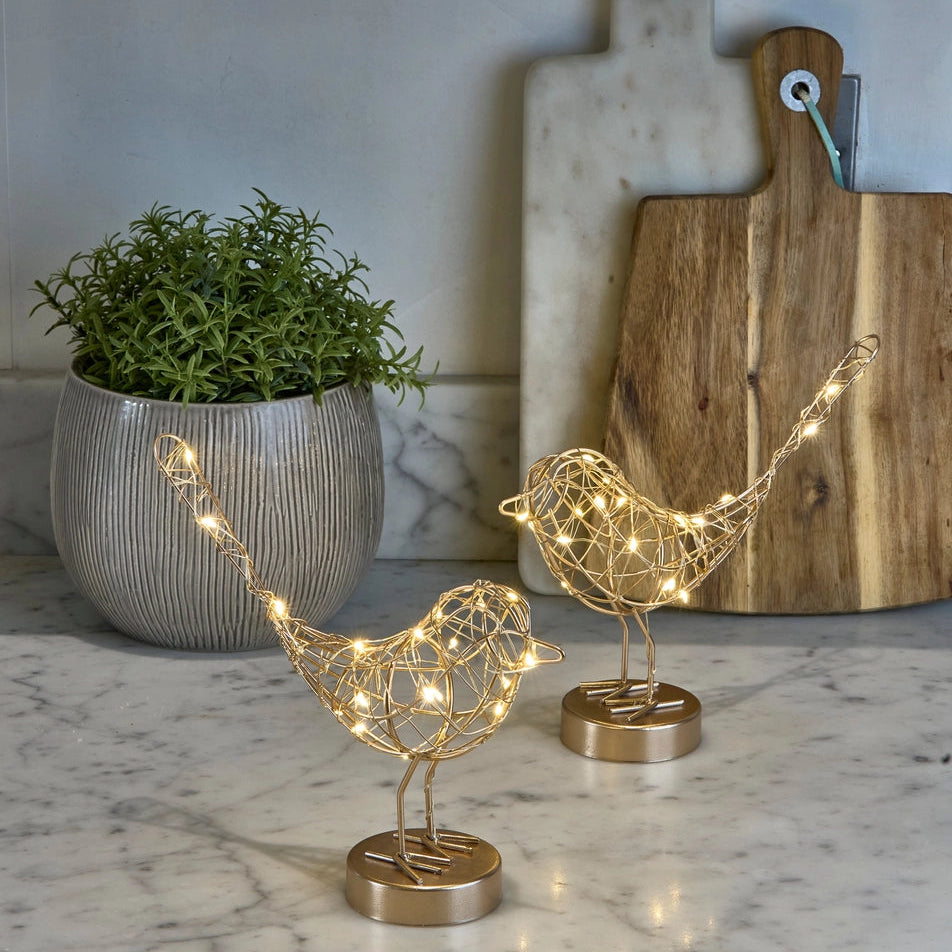 Decorative gold bird sculptures with string lights on a marble surface, with a plant and cutting board in the background.