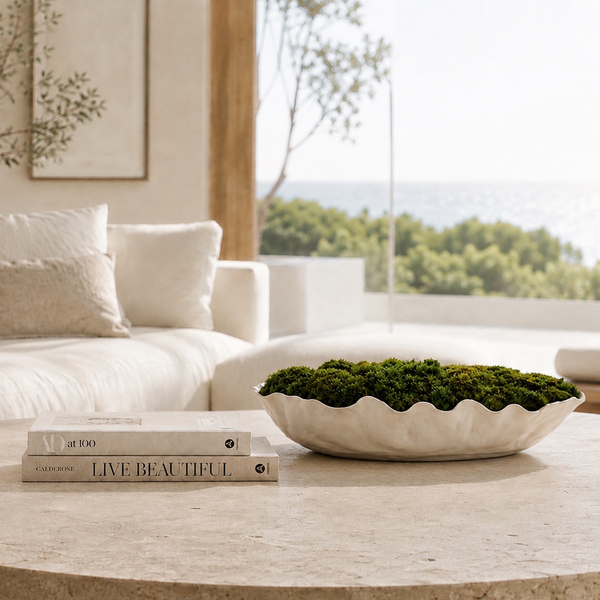 A styled interior scene with a white sofa, neutral pillows, a round stone coffee table, a lit candle, stacked books, and a white bowl filled with green moss. Large windows reveal trees and water outside.