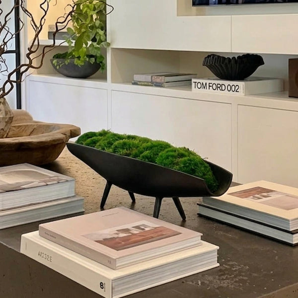 A black metal bowl-shaped planter filled with bright green moss, styled on a dark tabletop with books in a modern interior setting.