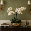 A decorative arrangement of white orchids in a gold bowl sits on a dark wood console table against an olive green textured wall with wall sconces and small decor objects.