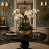 A black round pedestal table in a styled room, featuring a large gold bowl planter with white orchids, books, and a small gold lidded box. The background shows green patterned wallpaper, gold wall sconces, and a doorway.