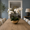 A styled dining room scene featuring a rustic wooden dining table with white upholstered chairs and a central gold bowl filled with white orchids, set against a slate blue wall and an open doorway.