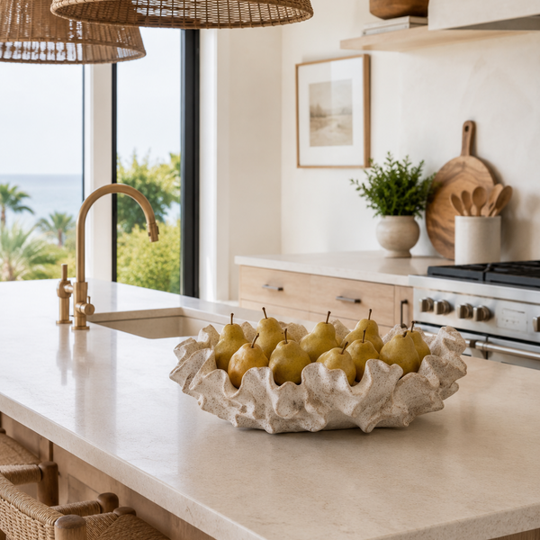 A modern kitchen scene with a sculptural textured bowl filled with pears on a light stone countertop. The kitchen features a brass faucet, a window overlooking greenery and the ocean, and warm wood accents.