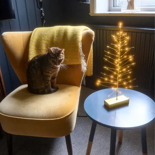 A small illuminated fiber-optic Christmas tree on a wooden base sits on a round blue side table next to a yellow armchair with a cat sitting on it in a dimly lit room.