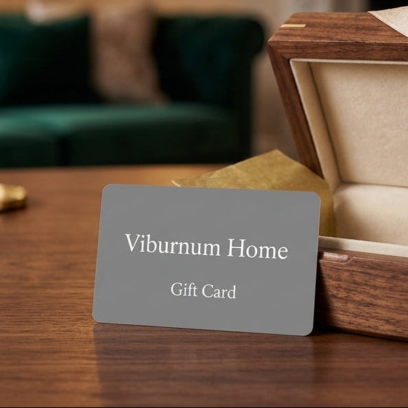 A gray gift card labeled 'Viburnum Home Gift Card' standing in front of an open wooden presentation box with a cream interior and ribbon, placed on a wooden table in a softly blurred living room setting.