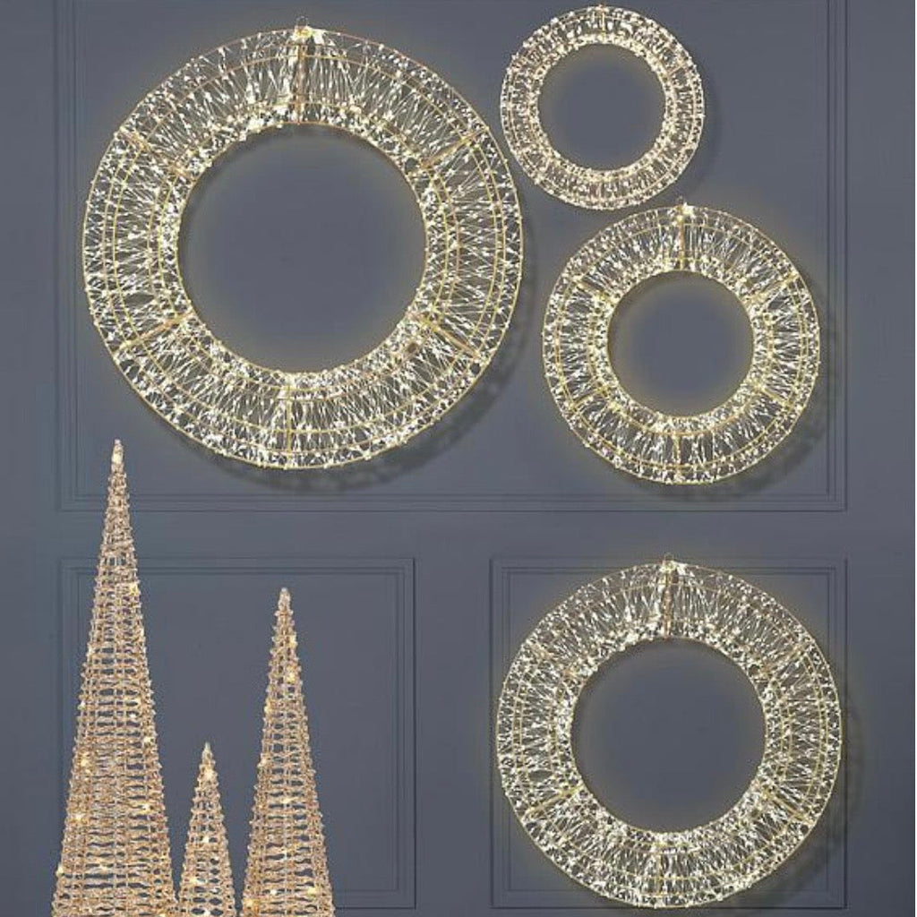 A set of illuminated wire decorative pieces: three circular wreath-shaped lights and three cone-shaped tree lights, shown lit against a dark gray paneled wall with a reflective floor.