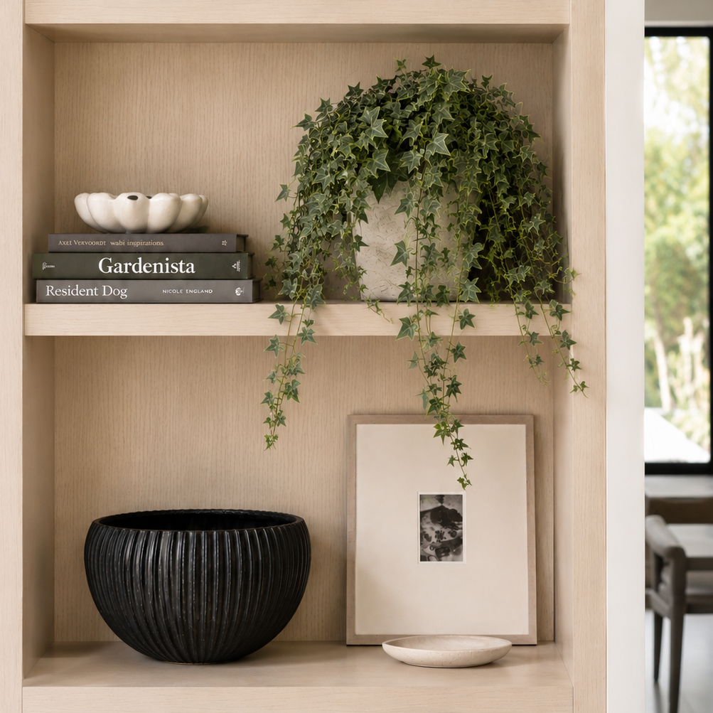 A light wood built-in shelving unit styled with home decor, including a white ceramic vase, a stone bowl, books, a potted trailing green plant, a black ribbed bowl, and a framed print.