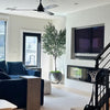 A modern living room interior with a dark blue sectional sofa, a light wood side table, a large potted olive tree, a built-in TV and fireplace, and a staircase with a small basketball hoop.