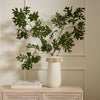 An artificial green leafy branch arrangement in a cream-colored ceramic vase with a pleated rim, styled on a light wood cabinet against a cream paneled wall.