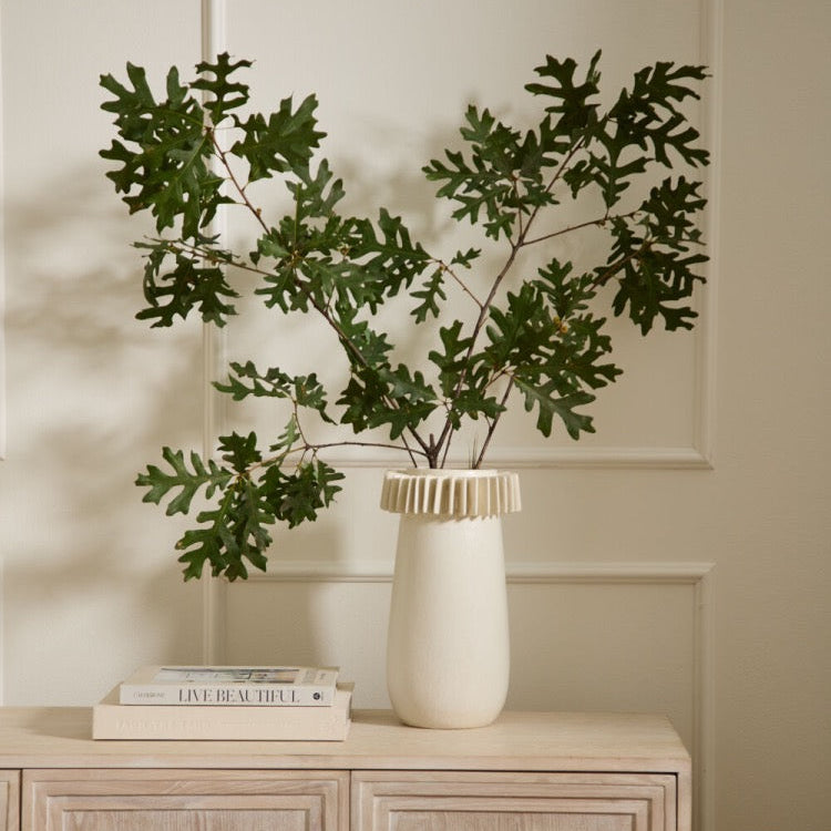 An artificial green leafy branch arrangement in a cream-colored ceramic vase with a pleated rim, styled on a light wood cabinet against a cream paneled wall.