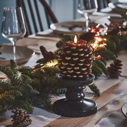A lit pinecone candle in a black pedestal holder on a dining table, surrounded by evergreen garland, pinecones, and warm string lights in a cozy indoor setting.