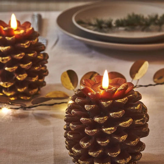 A decorative pinecone-shaped candle with a glowing flame sits on a beige tablecloth, with another similar candle and a plate in the softly blurred background.
