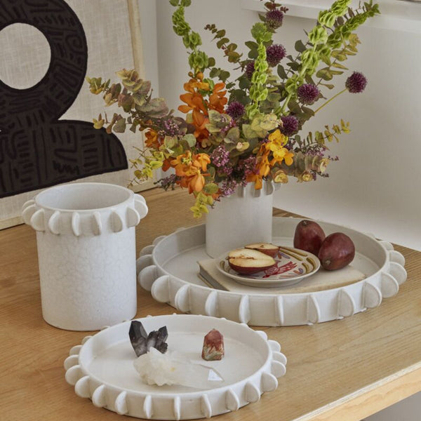 A white ceramic Reverie Tray with hand-applied decorative tabs, displayed on a wooden table with a floral arrangement, serving as a serving tray for a display of food and plants.