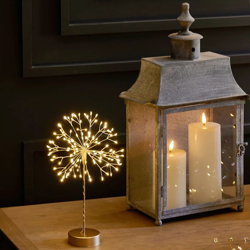 A decorative tabletop scene with a small gold wire light sculpture on a wooden table beside a rustic lantern containing two white candles. Warm string lights are draped across the table, and a dark paneled wall is in the background.