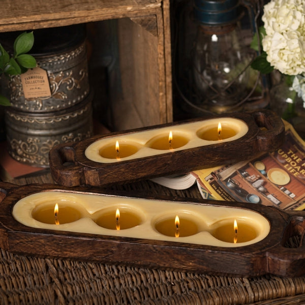 Two rustic wooden four-wick candle holders with lit candles, styled on a woven tray with vintage books and decorative containers in the background.