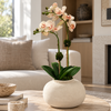 A faux Phalaenopsis orchid arrangement with pale blush pink flowers and green leaves, displayed in a round textured off-white ceramic pot on a round stone coffee table in a bright, modern living room.