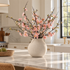 A ceramic vase filled with pink flowering branches arranged on a marble kitchen island, with a bright modern kitchen in the background.