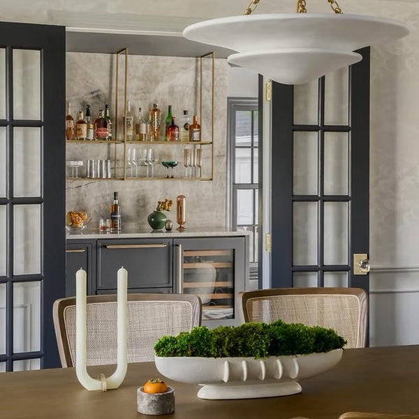 A modern dining room scene featuring a large white disc-shaped pendant light hanging from a gold chain over a wooden table. In the background, dark gray French doors frame an open doorway leading to a gray bar with gold shelving and bottles.