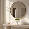 A large round wall mirror with a thin gold-toned frame hangs above a light stone console table in a bright, sunlit interior. A white pedestal bowl with green moss sits on the table, and the mirror reflects part of the room including a doorway and framed artwork.