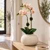 A potted artificial orchid with pale pink flowers and green leaves sits on a light wood console table in a bright, modern hallway. The orchid is housed in a textured off-white ceramic pot.