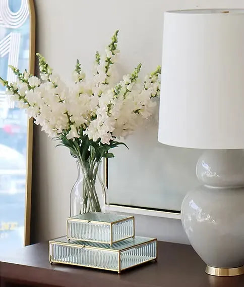 A styled interior scene showing a clear glass vase filled with white flowers on a dark wooden table, next to a white table lamp and a stack of mirrored glass boxes with gold trim.