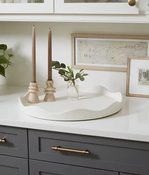 A round white ceramic tray with a wavy scalloped edge sits on a white countertop in a kitchen setting, styled with two tan taper candles in wooden holders and a small clear glass vase with greenery. Gray cabinets with brass handles are visible below, along with framed wall art.