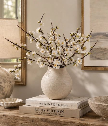 A textured cream ceramic vase filled with white blossoms on a stack of books on a wooden table, styled in a warm, elegant room setting.