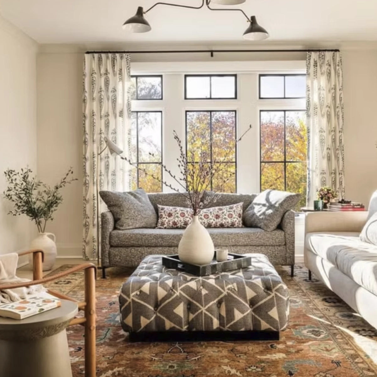 A styled living room featuring a gray patterned fabric sofa, a matching geometric ottoman, patterned curtains, and a large multi-pane window looking out to trees with autumn foliage.