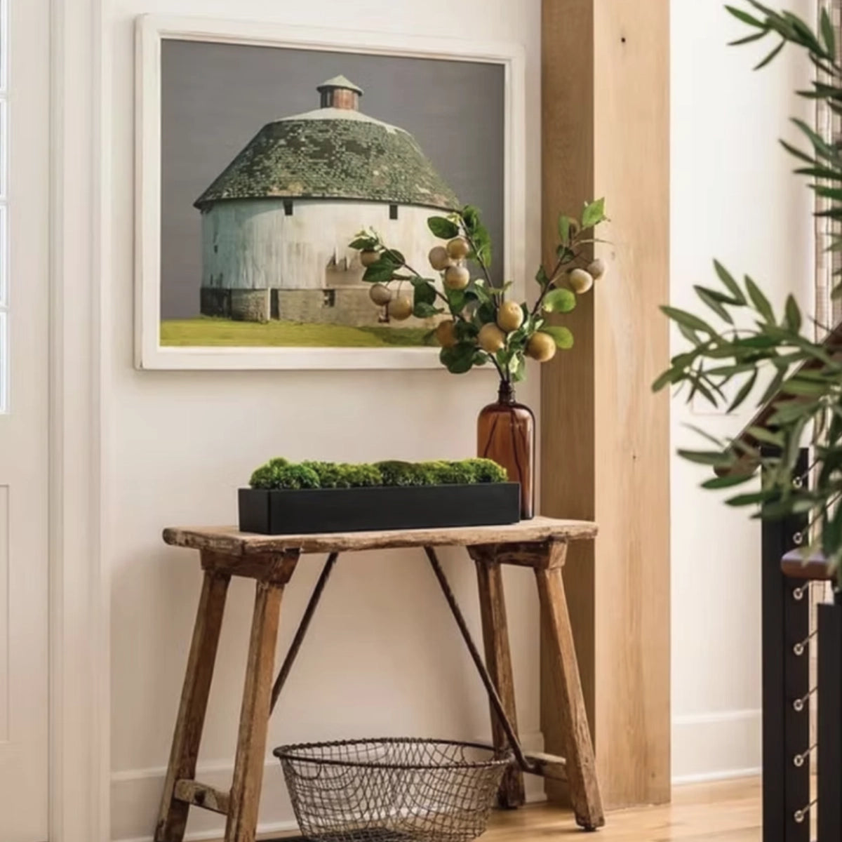 A framed art print depicting a rustic white barn with a mossy green conical roof, styled on a white wall above a rustic wooden table with a plant and a wire basket.