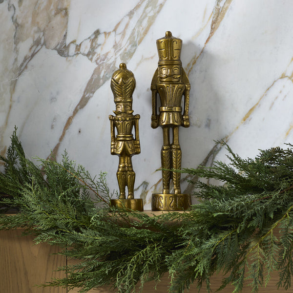 Two gold nutcrackers on a mantel with greenery against a marble wall.